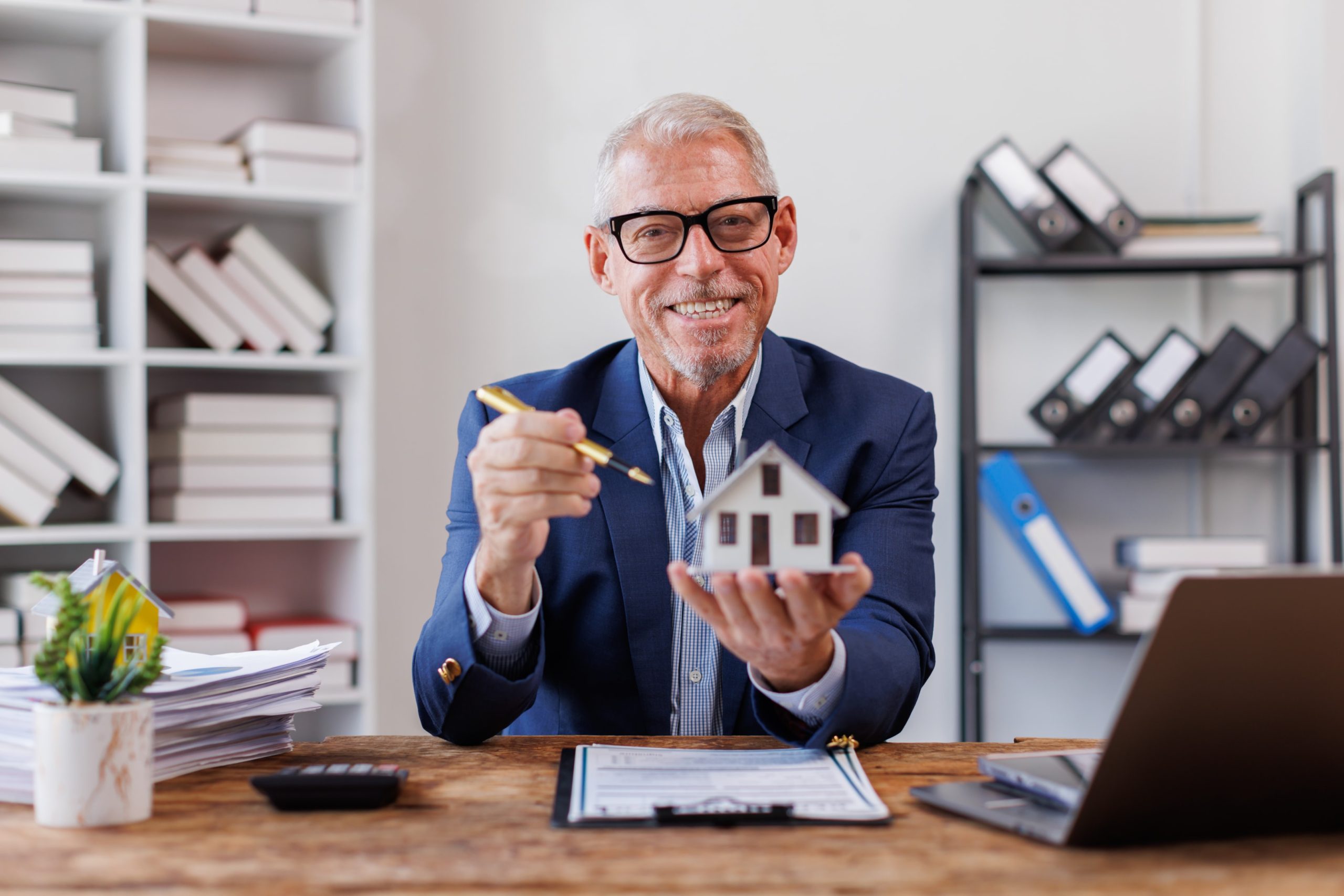 Smiling real estate or property lawyer holding a miniature house model at his desk, with documents, a laptop, and office files in the background.